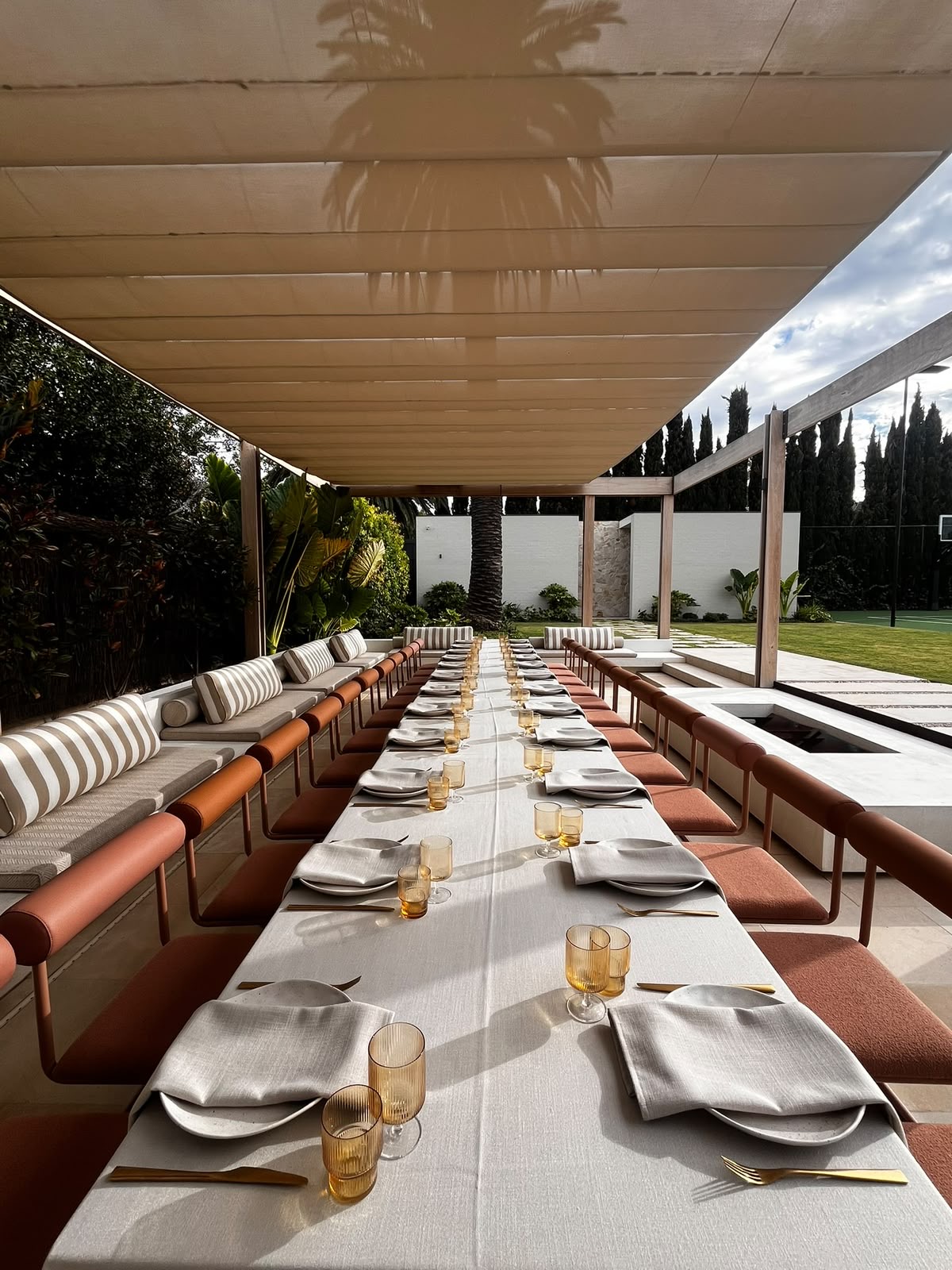 A long outdoor dining table set with linen, folded napkins and amber glassware beneath a slatted pergola