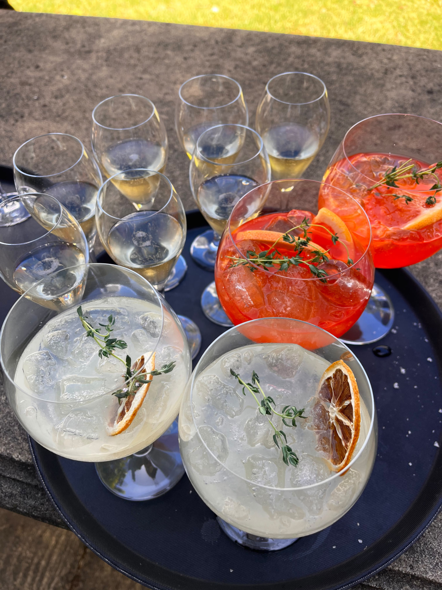 A black tray of cocktails and sparkling wines garnished with citrus wheels and thyme, resting on a stone ledge in sunlight