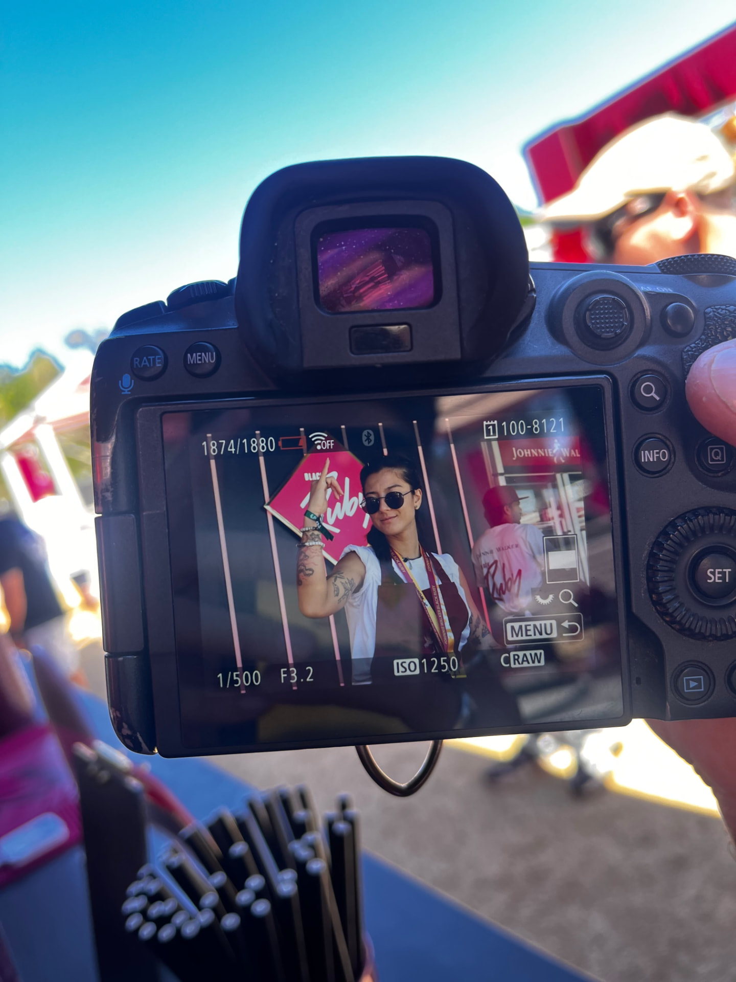 A camera held between guests, its screen showing a smiling woman in sunglasses at an event backdrop